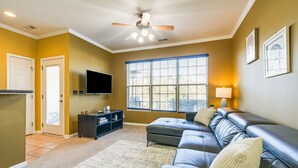 A living room with a dark leather sofa, a flat-screen TV, a ceiling fan, and a large window with blinds. - Spacious Retreat Near RDU ~ Pool, BBQ, Fast Wi-Fi & Easy Self Check-In (Morrisville)