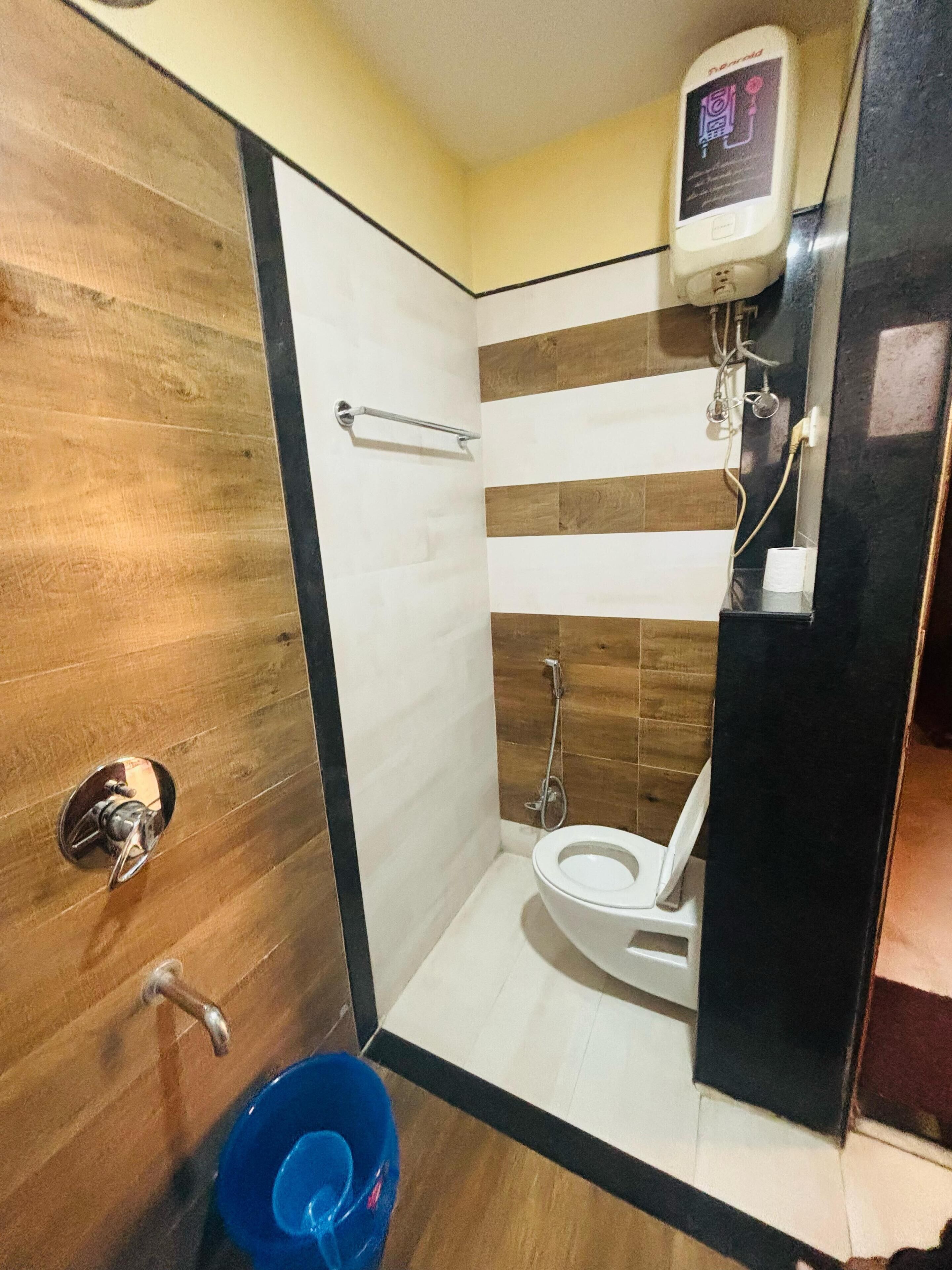 A small bathroom with a wooden floor, a white toilet, a blue bucket, and a hand dryer.