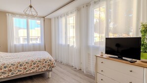 Ein Schlafzimmer mit einem Bett, einem Fernseher auf einer Kommode und großen Fenstern mit Vorhängen.