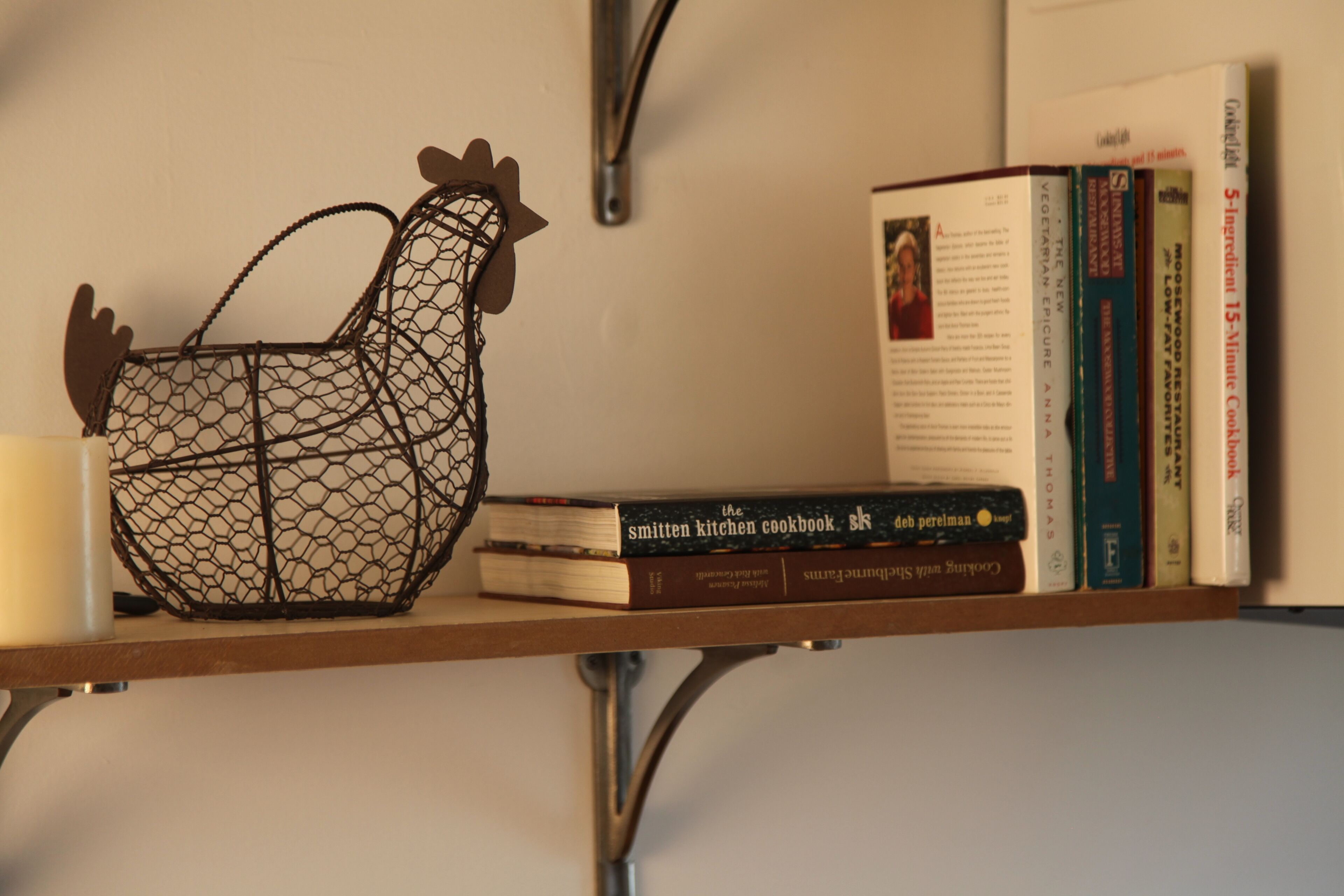 A shelf with a metal chicken-shaped container, several cookbooks, and a candle.