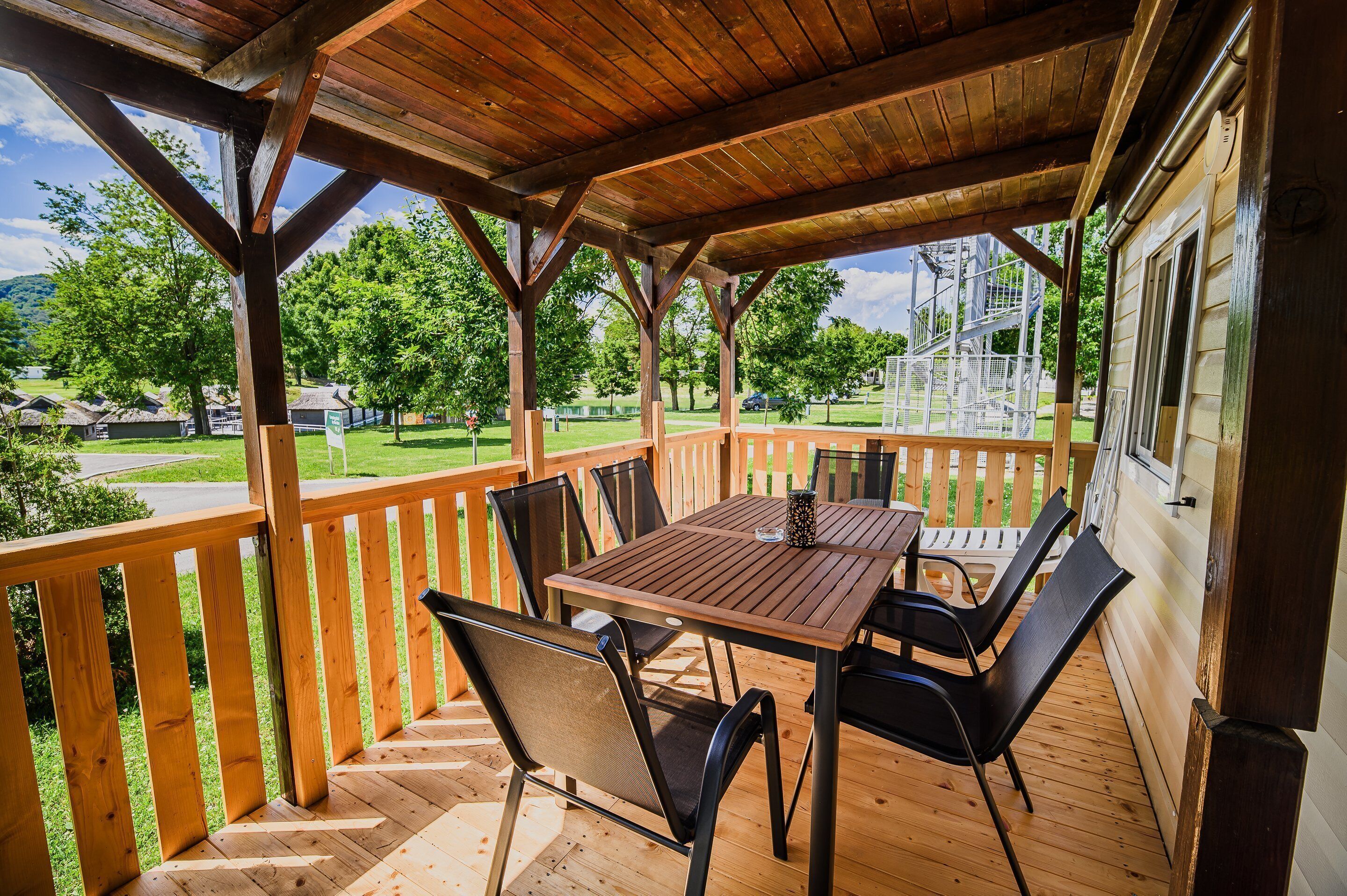 A covered outdoor patio with a wooden table and chairs, a wooden railing, and a view of a park and buildings in the background.