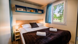 A neatly made bed with a dark brown quilt, a wooden headboard, and a bedside table with a lamp. There are two blue shelves above the bed, and a window with blue curtains on the right. - Waterside Retreat Mobile Homes With Thermal Riviera Tickets (Brežice)