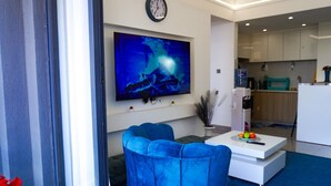 A modern living room with a blue sofa, a flat-screen TV, and a small dining table with fruit. - Lux Suites Riara One Residency Kilimani (Nairobi)