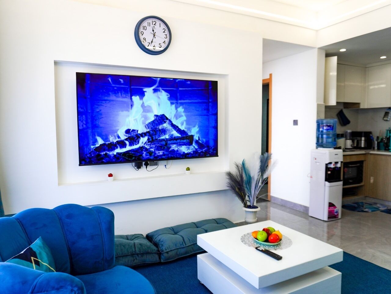 Una sala de estar moderna con un televisor de pantalla plana grande, un sofá azul, una mesa de centro blanca con frutas y una cocina al fondo.