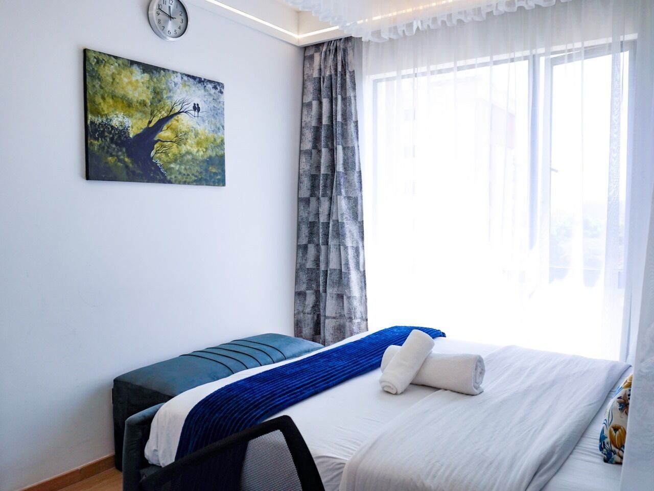 A hotel room with a bed, a painting of a tree, a window with curtains, and a clock on the wall.