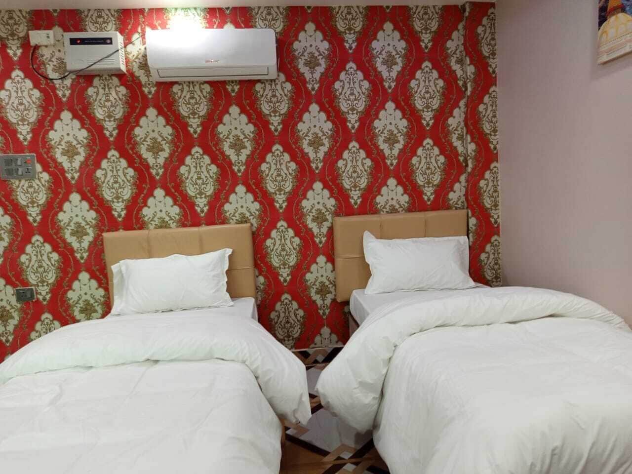 Habitación de hotel con dos camas, un papel tapiz rojo estampado, un aire acondicionado y un enchufe en la pared.