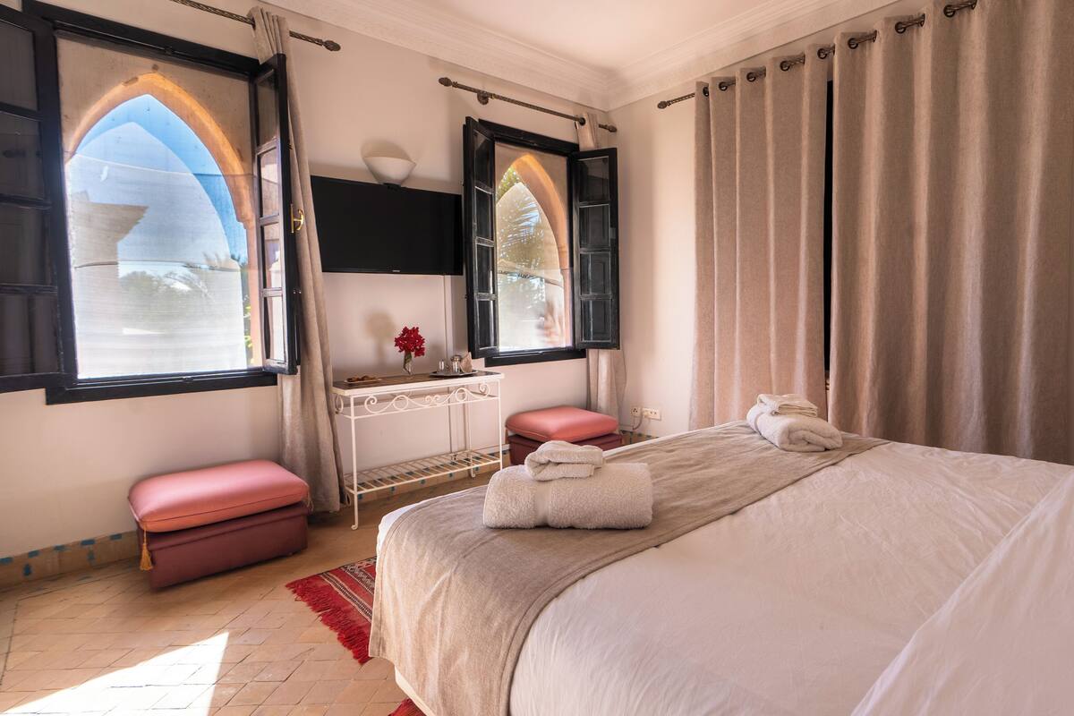 A bedroom with a bed, a vanity with a mirror, a window with curtains, and a red bench.