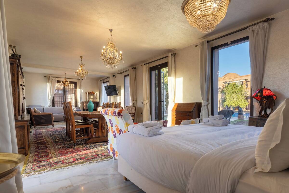A hotel room with a large bed, a dining table, a TV, and a view of the outside.