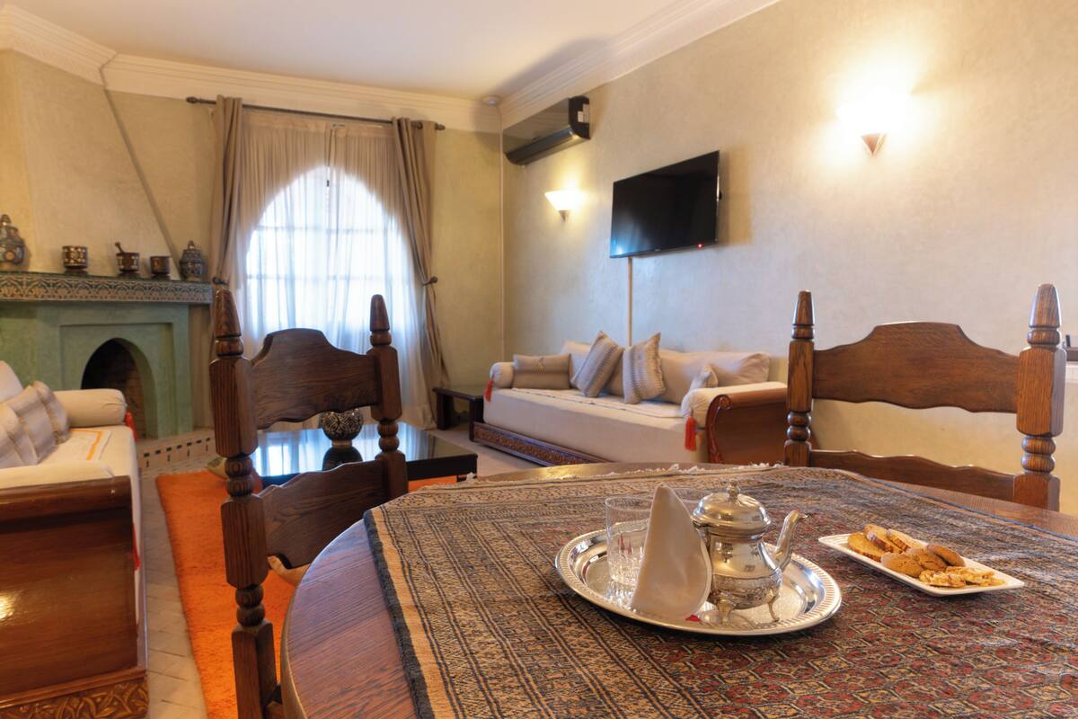 A hotel room with a bed, a sofa, a dining table with a tray of cookies, a fireplace, and a television mounted on the wall.