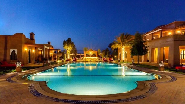 A large, well-lit swimming pool with a tiled deck surrounding it, set against a backdrop of buildings and palm trees. - VILLA THAIFA (Marrakech)