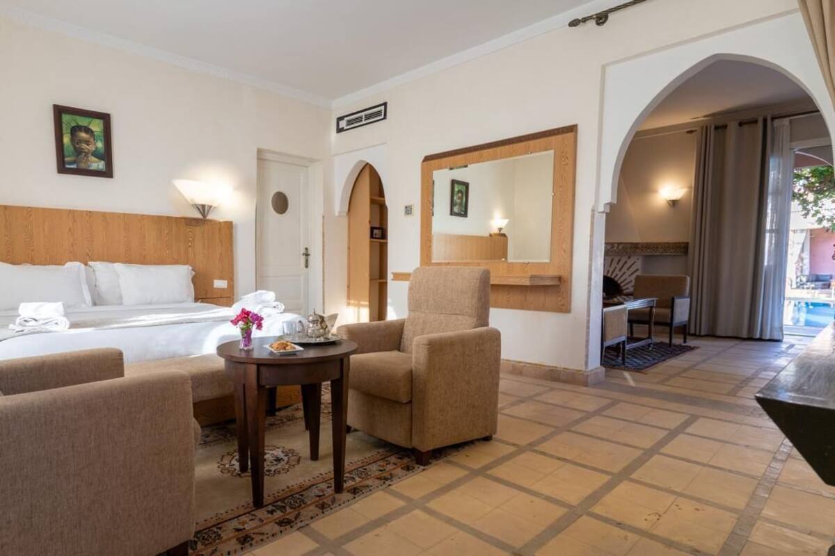 A hotel room with a bed, two armchairs, a small table, a mirror, and a door leading to a balcony.
