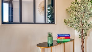 Ein modernes Interieur mit einem Fenster, einem Wandkunstwerk, einer Topfpflanze und einer Stapel bunter Bücher auf einer Konsole.