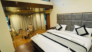 A spacious bedroom with a large bed, a separate seating area with a chair and table, and a view of the outdoors through large windows. - Thiksay Organic Resort and Retreat (Leh)