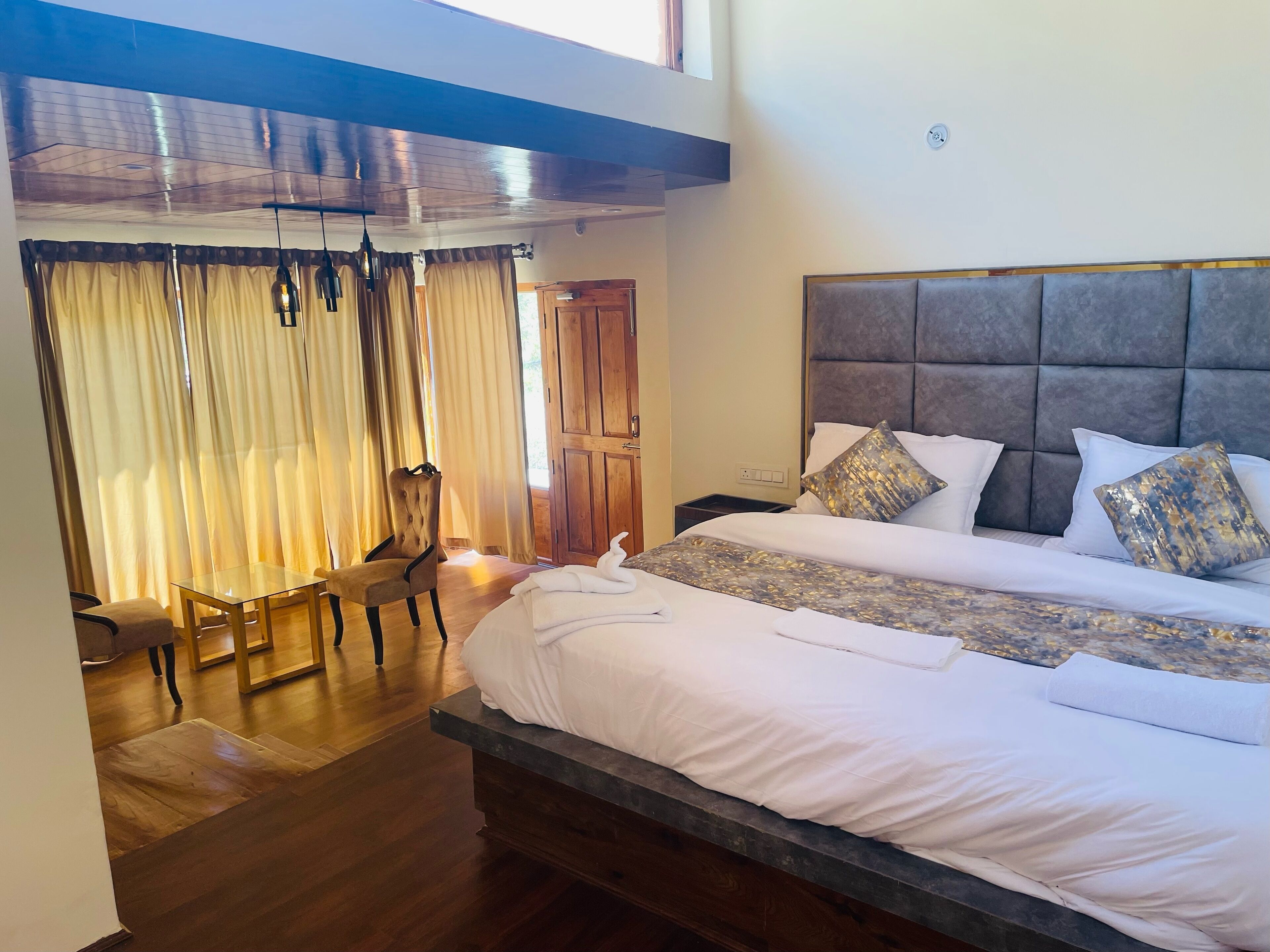 Une chambre d’hôtel avec un grand lit, une tête de lit en bois, un coin repas avec une table en verre et des rideaux, et une vue sur l’extérieur.