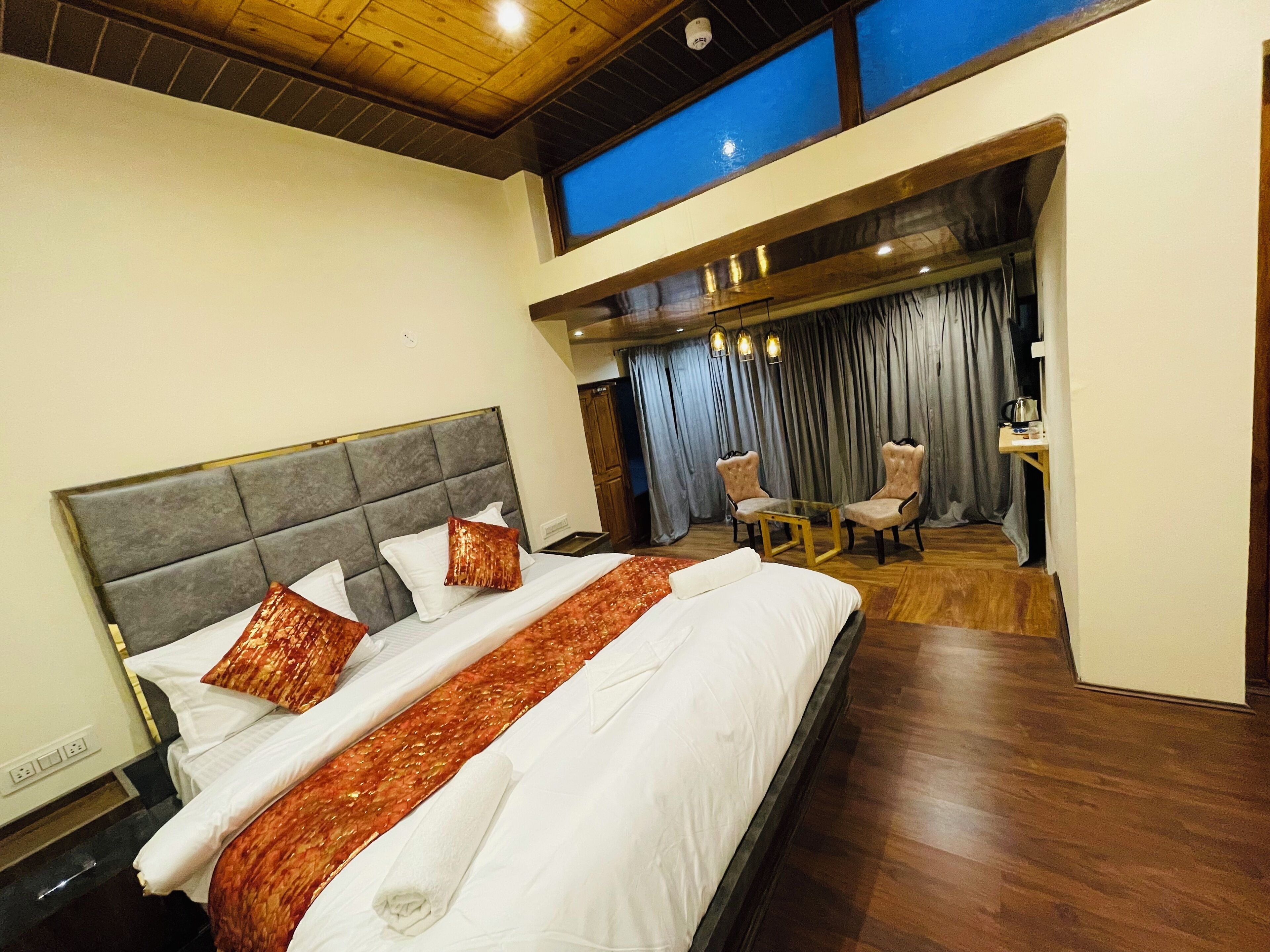 A hotel room with a large bed, a small table with chairs, and a view of the sky through a window.