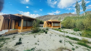 Two traditional-style buildings with wooden facades and large windows, set in a desert landscape with sparse vegetation and mountains in the background. - Thiksay Organic Resort and Retreat (Leh)