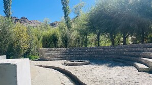 A stone-walled outdoor area with a circular stone fire pit, surrounded by a paved walkway and greenery. - Thiksay Organic Resort and Retreat (Leh)