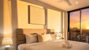 A hotel room with a bed, a ceiling fan, a nightstand with a lamp, a window with a sunset view, and a framed artwork on the wall. - Sky-High 2BR|Spectacular Views|Free Parking! (South Brisbane)