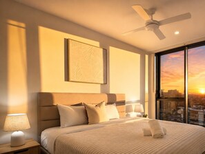 A hotel room with a bed, a ceiling fan, a nightstand with a lamp, a window with a sunset view, and a framed artwork on the wall.