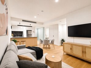A modern living room with a sofa, a dining table with chairs, a kitchen area, and a television mounted on the wall.