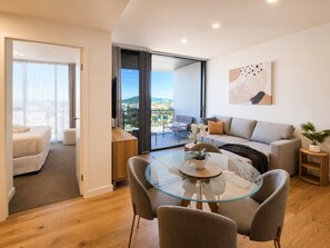 A modern living room with a glass dining table, a sofa, and a view of a cityscape through large windows.