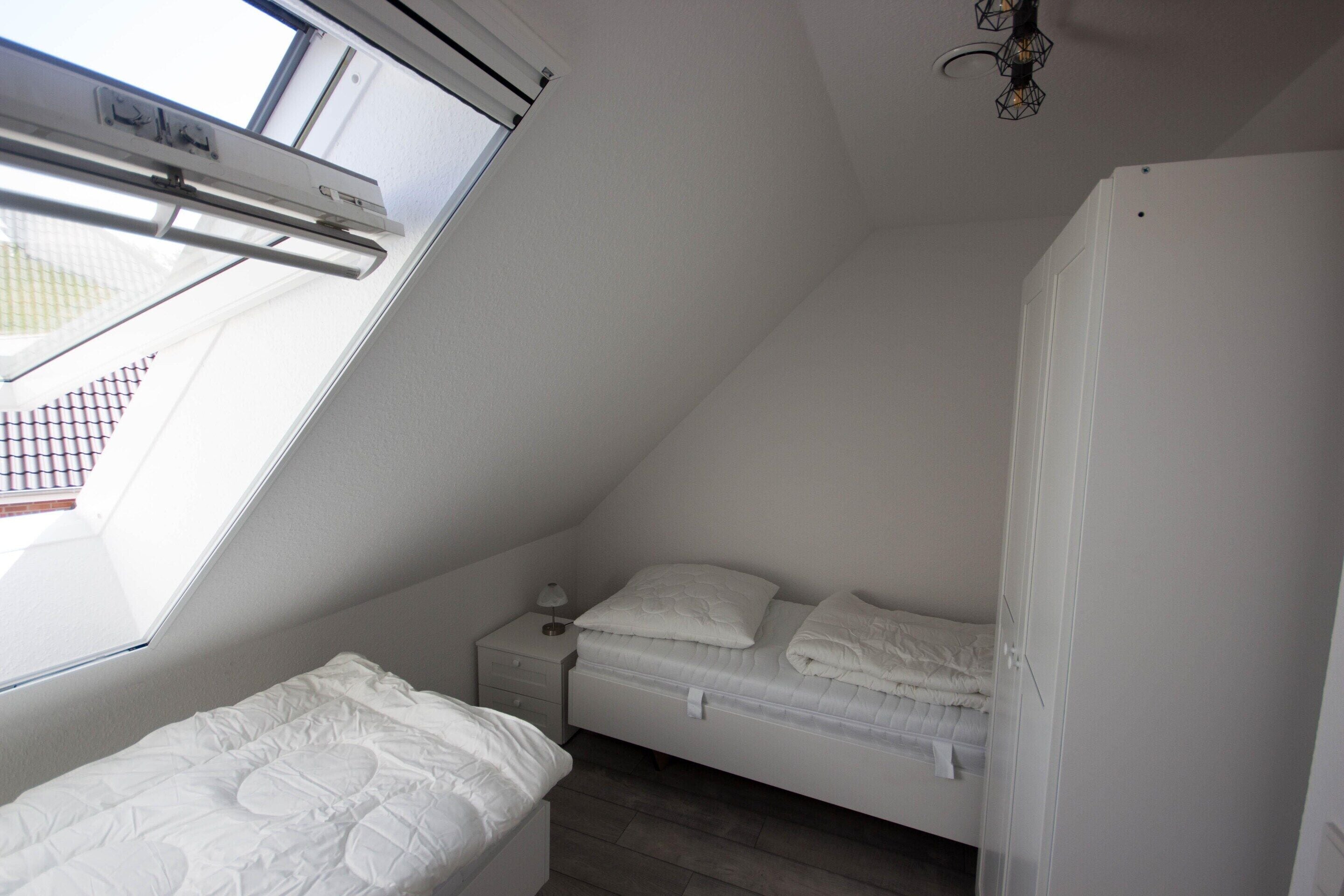 Una habitación pequeña y blanca tipo residencia de estudiantes, con tragaluz, cama, mesita de noche y armario.
