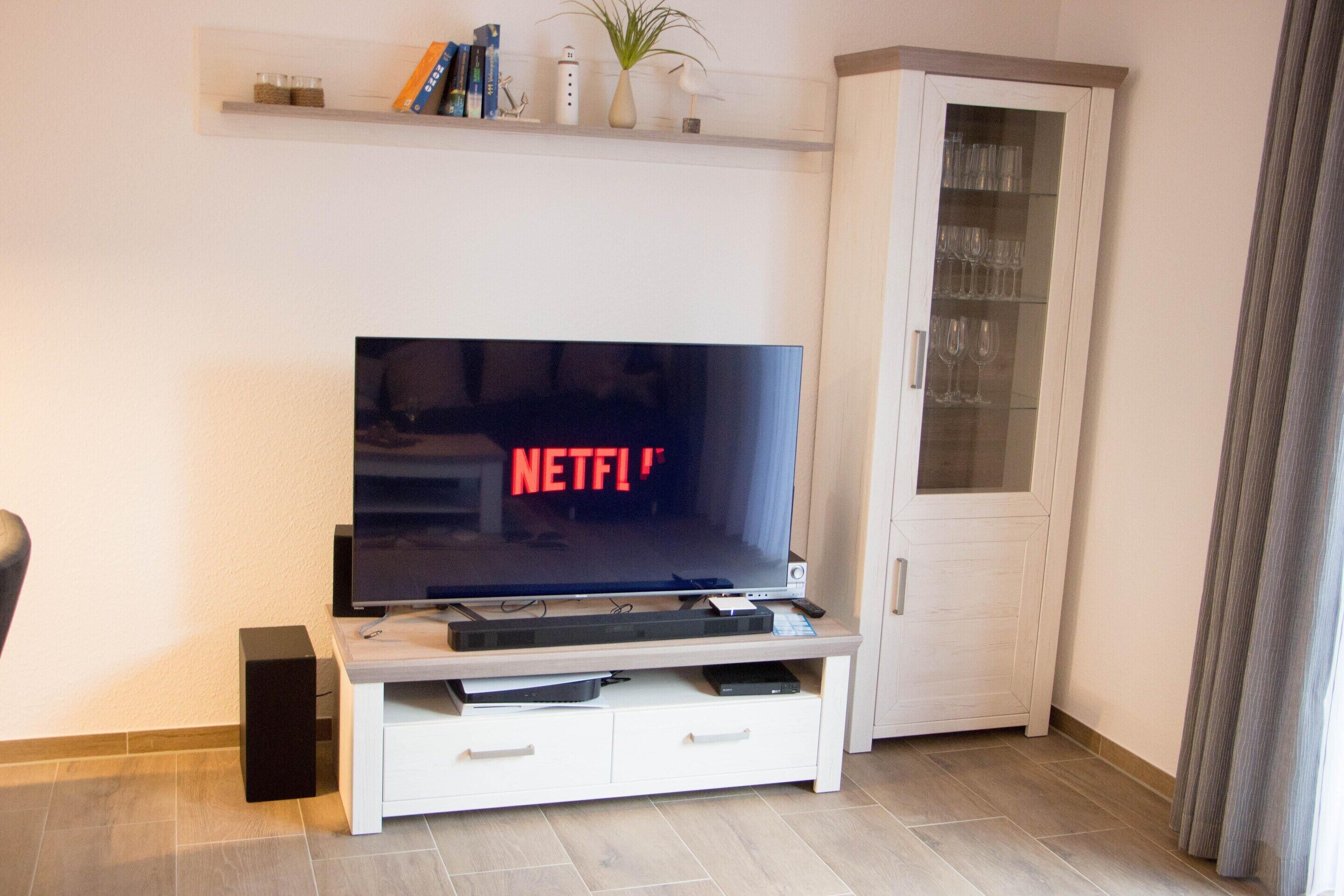 A living room with a flat-screen TV on a white entertainment unit, a cabinet with glass doors, and a shelf with books and decorative items.