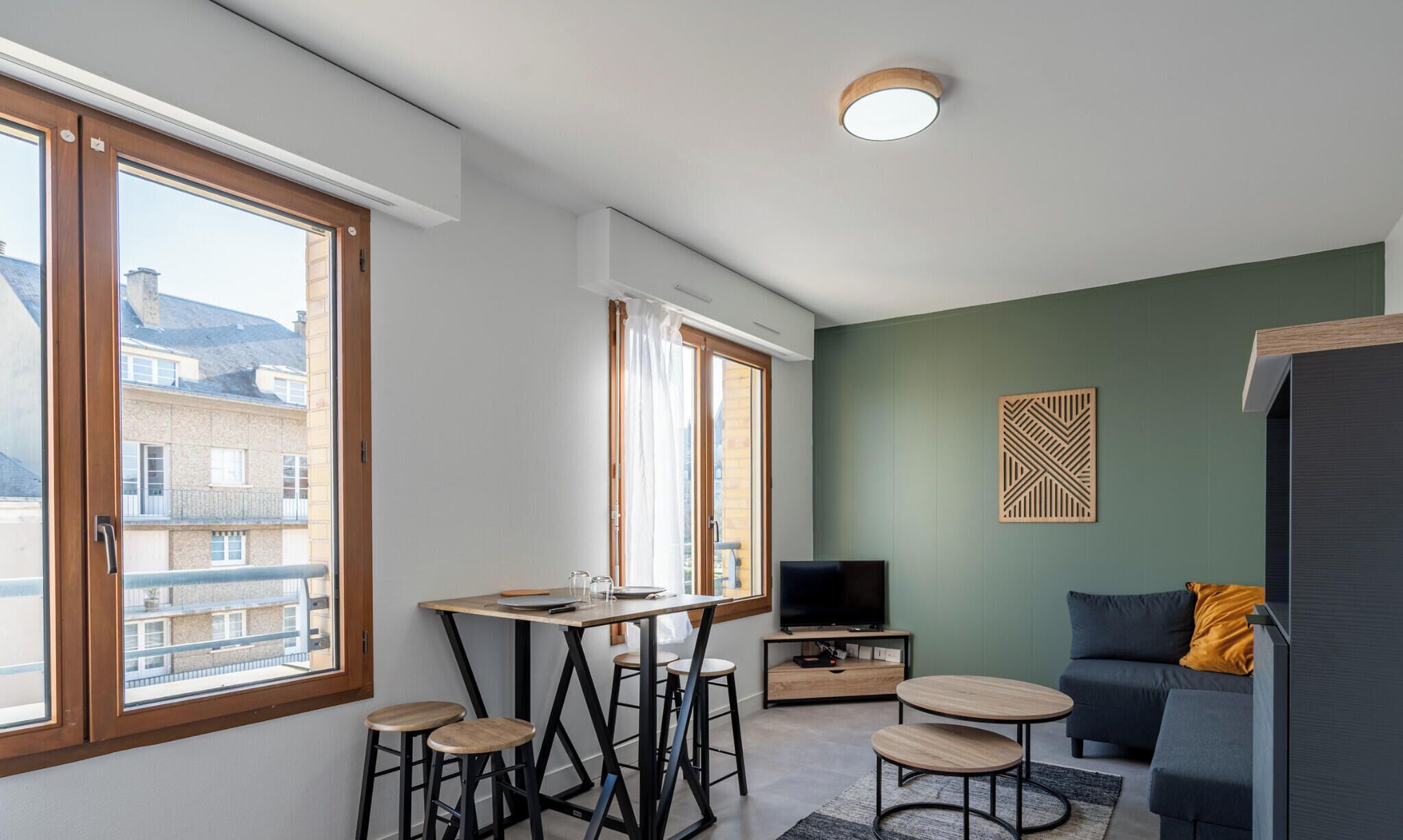 Een modern appartement met grote ramen, een eethoek, een woonkamer met een bank en televisie, en een decoratieve wand met een patroon.