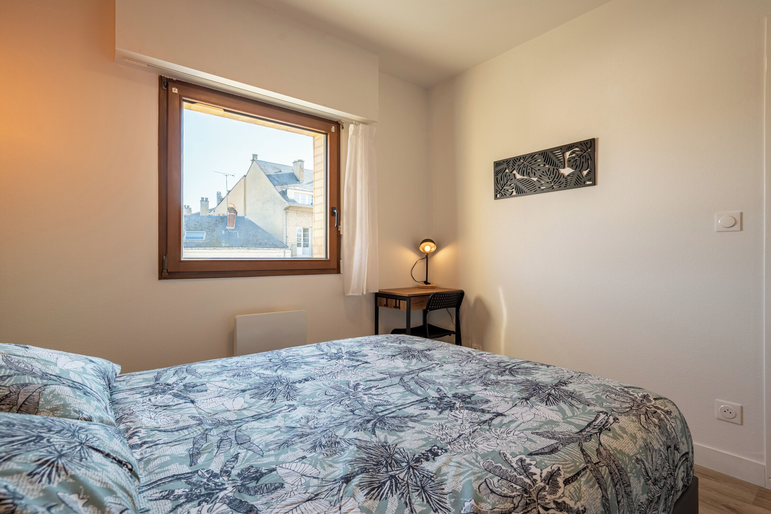 A bedroom with a bed, a window, a desk, and a wall-mounted light fixture.