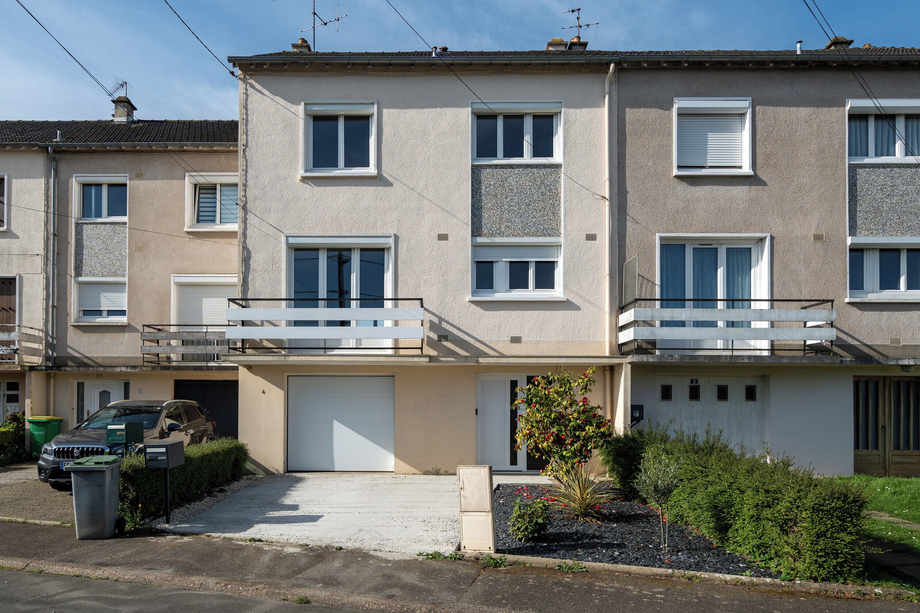 Un immeuble résidentiel à plusieurs étages, doté d’un garage, avec une voiture garée devant et un petit jardin agrémenté d’arbustes et d’un arbre.