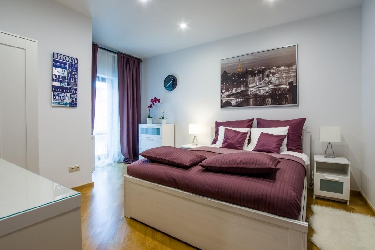 Un dormitorio con cama, mesita de noche, lámpara, reloj, ventana con cortinas y un cuadro en la pared.