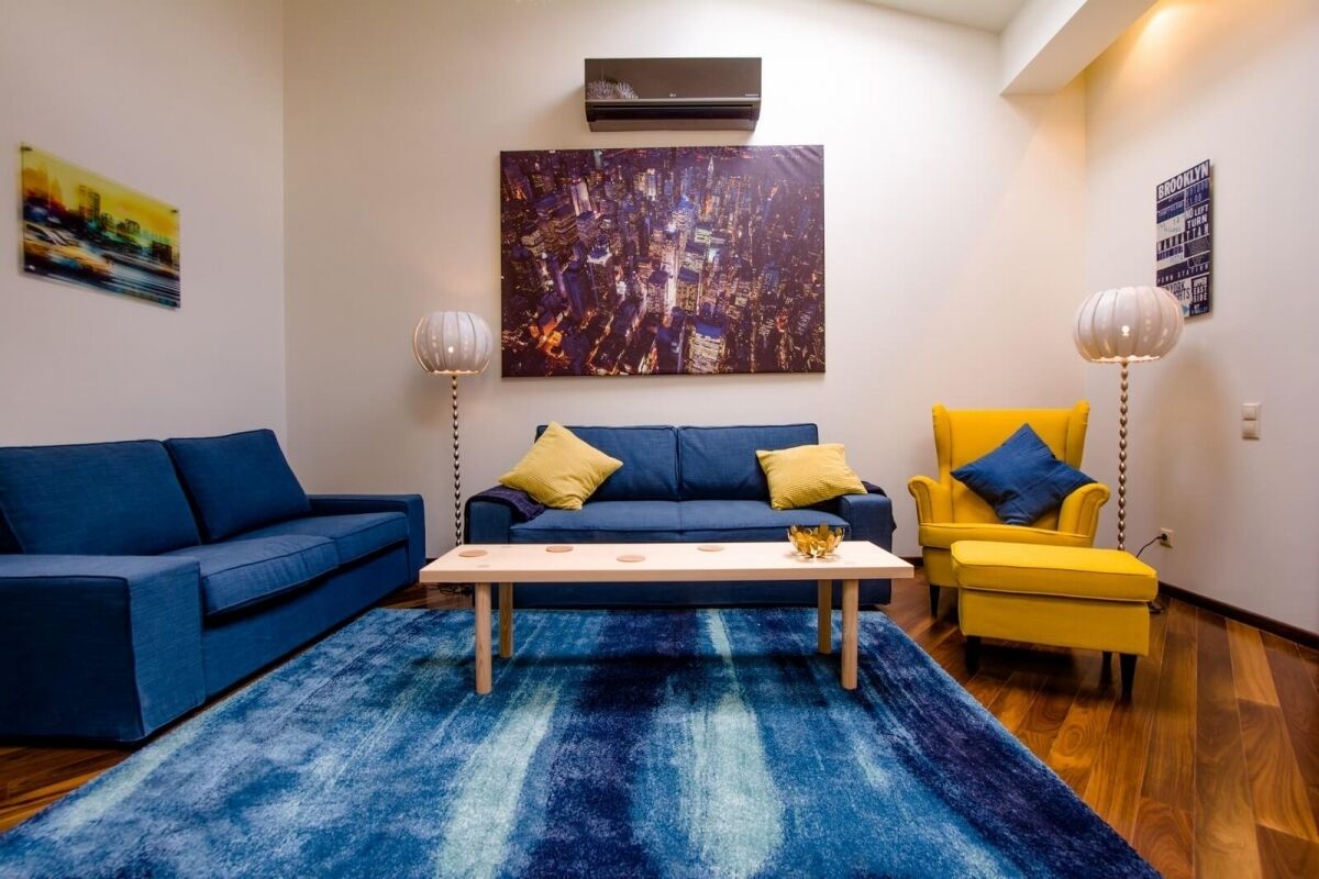 A living room with a blue sofa, a yellow armchair, and a coffee table. There is a cityscape painting on the wall and an air conditioning unit above the sofa.