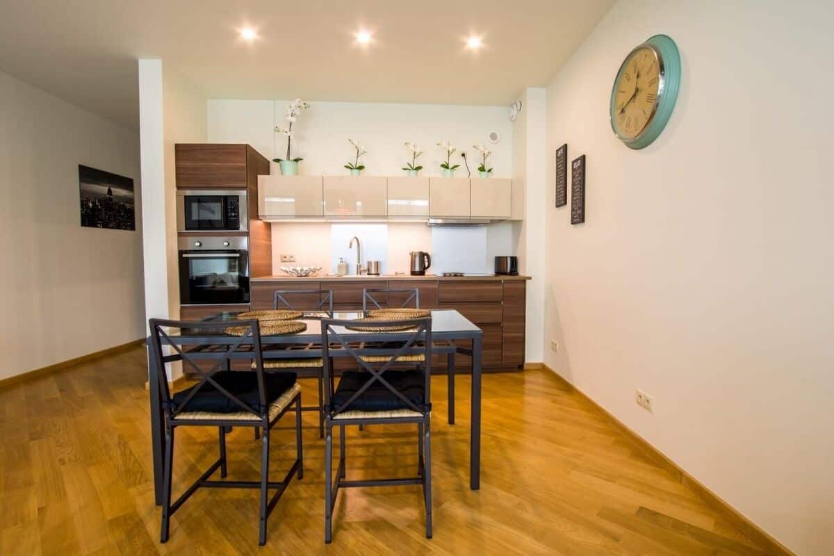 Una cocina moderna con electrodomésticos integrados, un comedor con una mesa y sillas de madera, y un reloj en la pared.