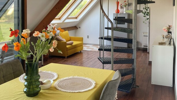 Un loft moderne doté d’un espace repas agrémenté d’une nappe jaune, d’un vase en verre contenant des fleurs et d’un plafond en bois avec des poutres apparentes.