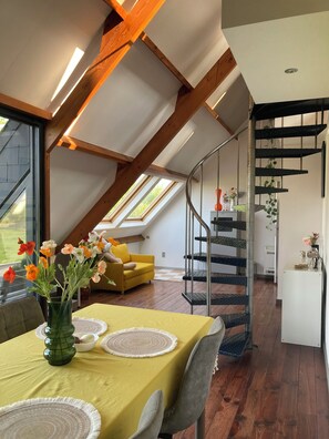 Un loft moderne doté d’un espace repas agrémenté d’une nappe jaune, d’un vase en verre contenant des fleurs et d’un plafond en bois avec des poutres apparentes.