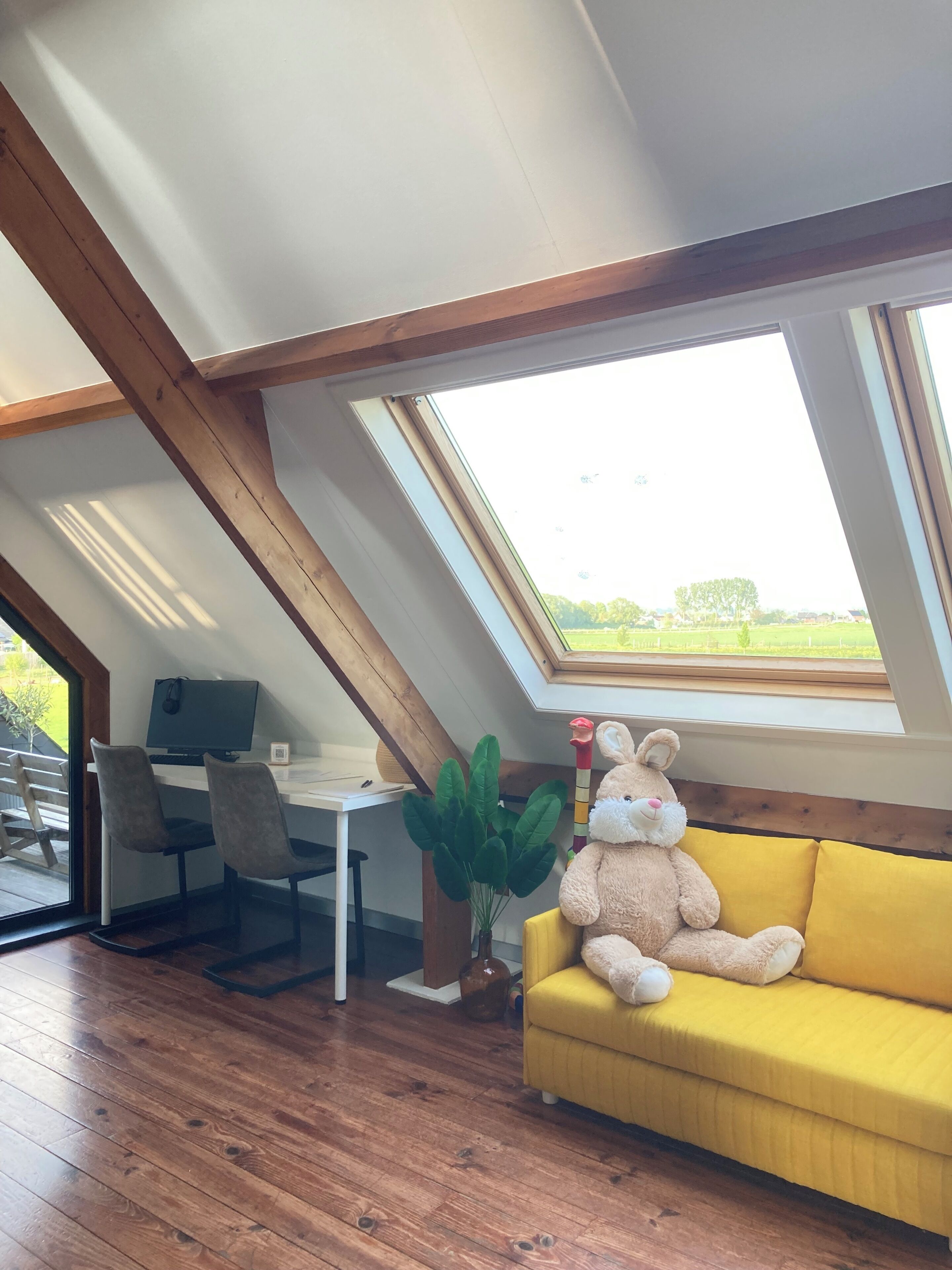 Una habitación con techo inclinado, un sofá amarillo, un ventanal grande, un escritorio con una computadora y una planta.