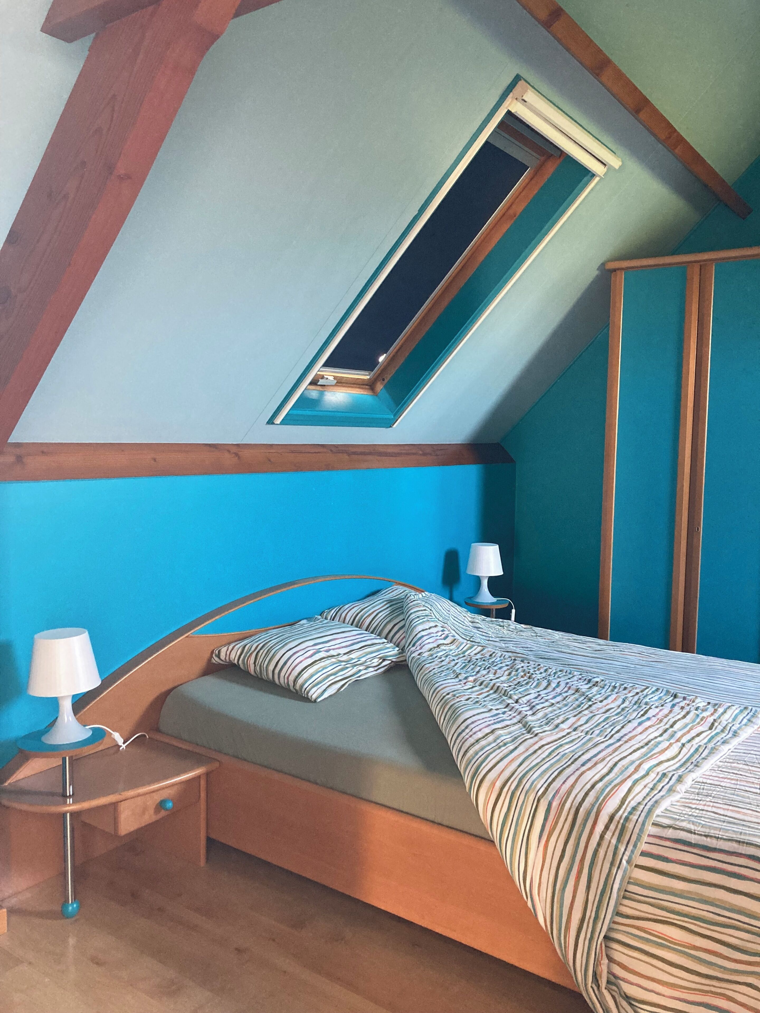 Ein Schlafzimmer mit geneigter Decke, einem Oberlicht, einem Bett mit gestreifter Bettwäsche, einem Nachttisch und einer Lampe.