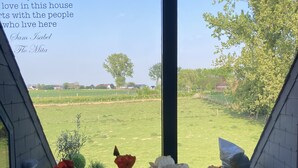 Vue depuis une pièce dotée d’une grande fenêtre donnant sur un champ verdoyant et des arbres. On peut lire une citation sur la fenêtre, et un vase rempli de fleurs est posé sur le rebord de la fenêtre.