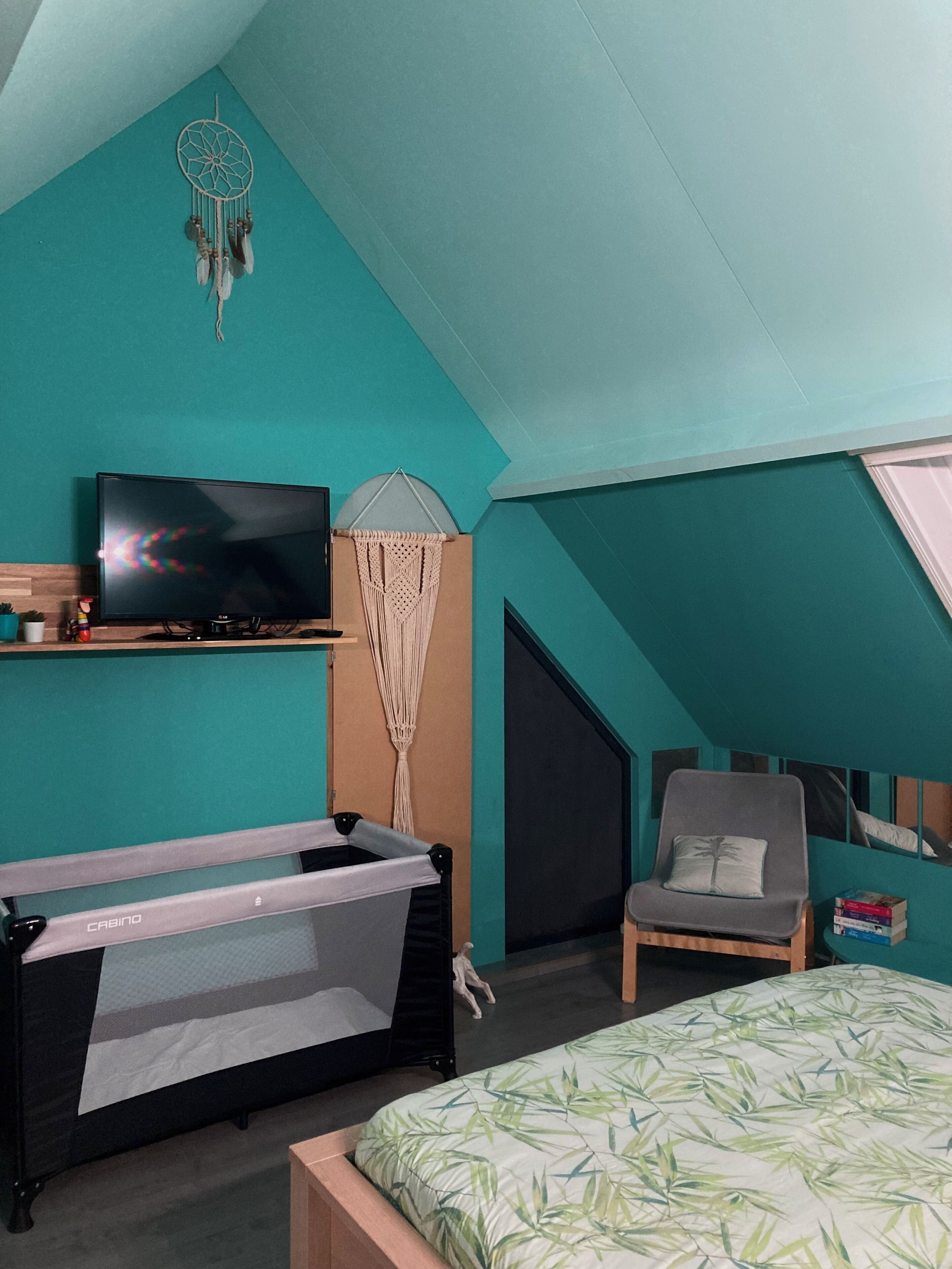 Une chambre avec des murs de couleur turquoise, un lit recouvert d’une courtepointe à motifs de feuilles vertes, un téléviseur posé sur une étagère en bois, une décoration murale en macramé, une chaise avec un coussin et un berceau.