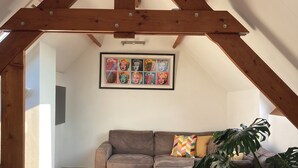 Un salon chaleureux de style loft, avec un plafond en bois, un canapé d’angle gris, une grande plante et une reproduction d’une œuvre de pop art encadrée au mur.