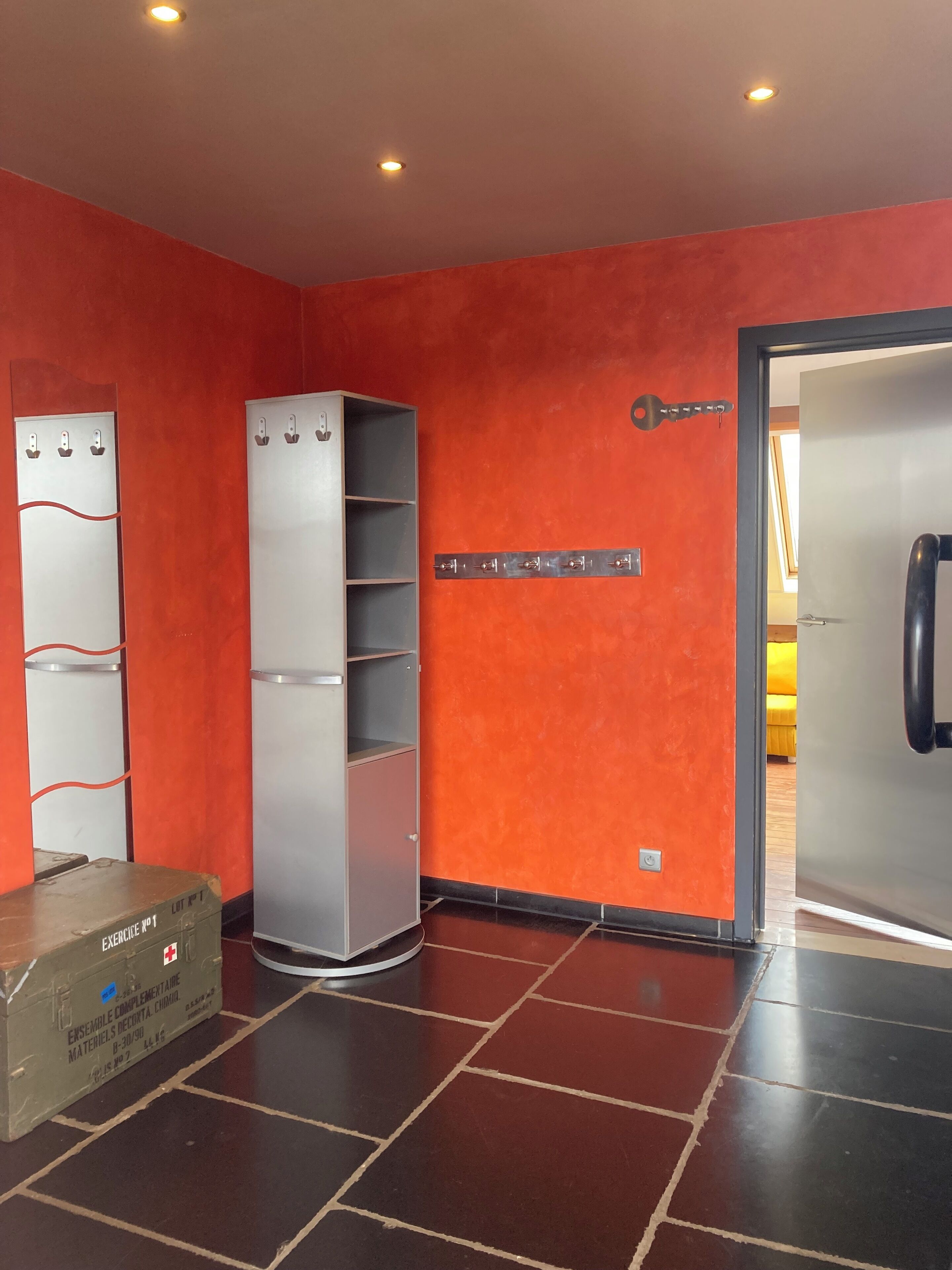 Una stanza con un muro rosso, un armadio grande, un mobiletto con ganci e una porta con maniglia.