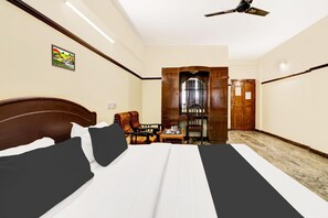 A hotel room with a large bed, a wooden wardrobe, a chair, a desk, and a ceiling fan.