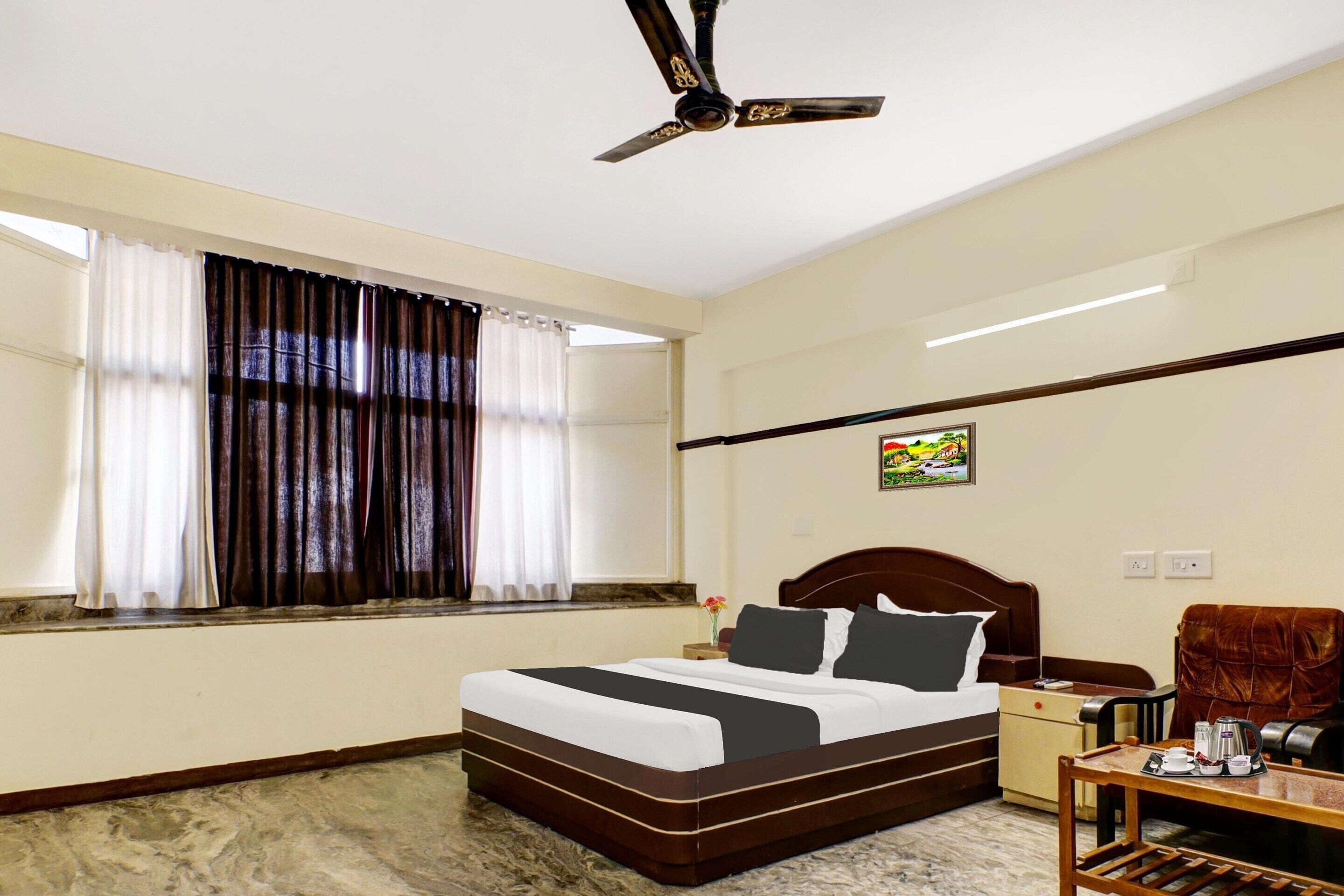 A hotel room with a bed, a nightstand, a chair, a table with a kettle, and a ceiling fan.
