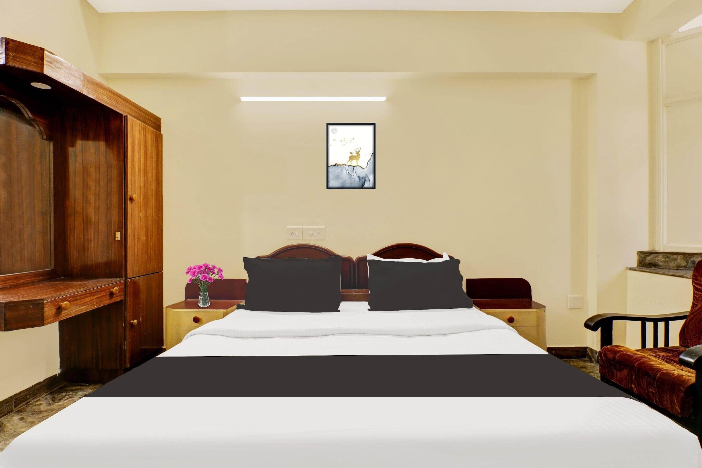 A hotel room with a bed, a wooden wardrobe, a nightstand with a vase of flowers, and a framed picture on the wall.