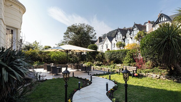 A garden patio with outdoor furniture, a curved pathway, and a large umbrella. - Annan Hotel (Llandudno)