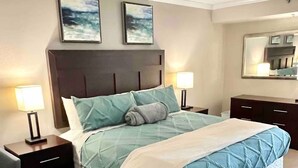 A neatly made bed with a quilted headboard, two bedside tables with lamps, a gray armchair, and two framed pictures on the wall. - Oceanfront Double-Room; Oceanview, Privacy & Space (Myrtle Beach)
