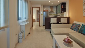 Un salon moderne avec un canapé et une table basse, un coin cuisine équipé d’un réfrigérateur et de placards, et un couloir menant à une salle à manger.