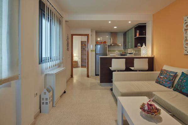 Una sala de estar moderna con un sofá y una mesa de centro, una zona de cocina con refrigerador y alacenas, y un pasillo que conduce a un comedor.