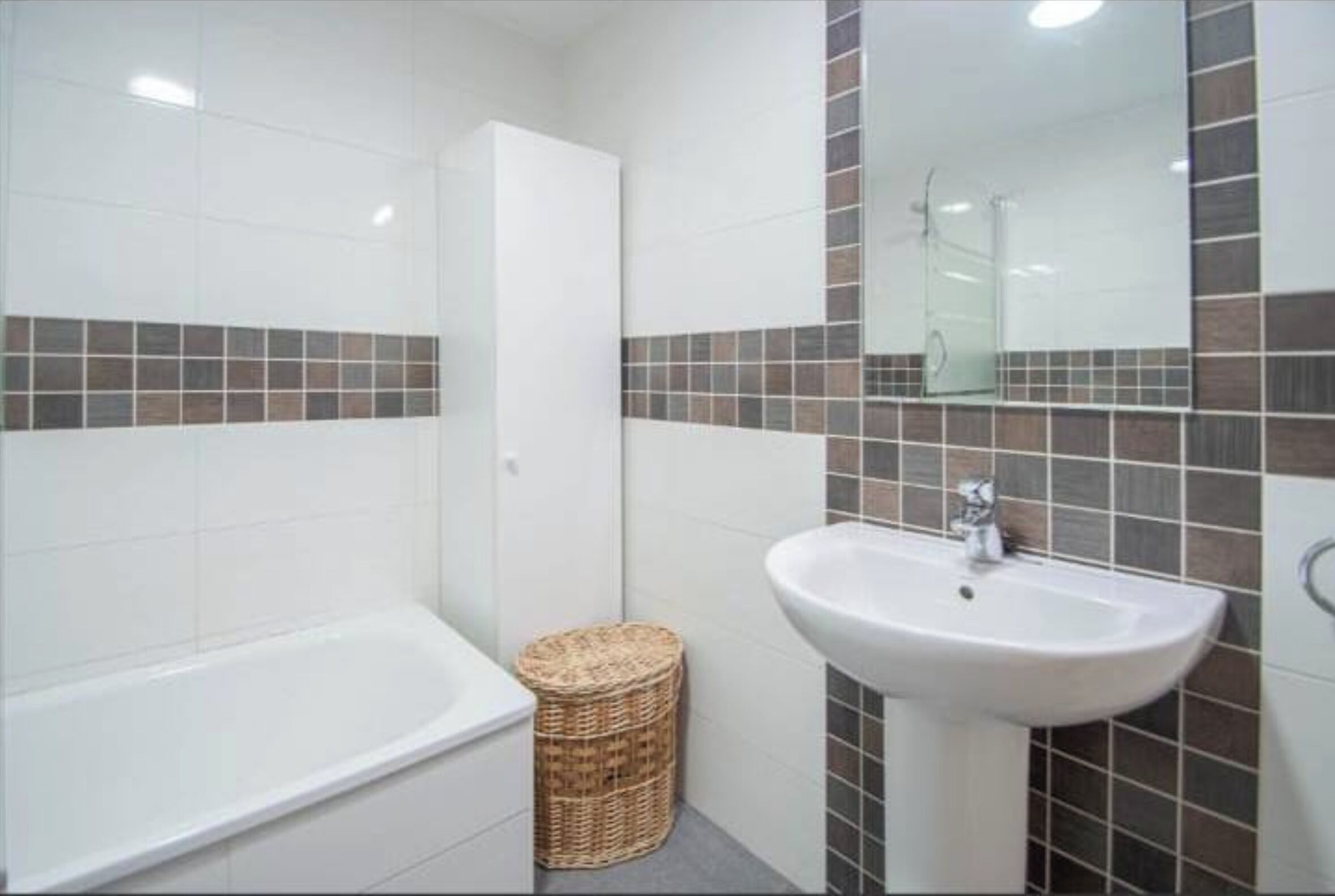 Un baño con una bañera blanca, un lavamanos blanco y un espejo sobre el lavamanos. Hay una cesta de mimbre en el suelo.