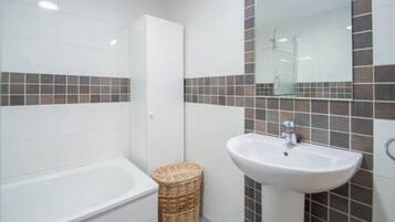 Un baño con una bañera blanca, un lavamanos blanco y un espejo sobre el lavamanos. Hay una cesta de mimbre en el suelo.